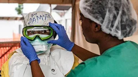 Getty Images A person in PPE and full head covering is having their goggles adjusted by someone, with blue protective gloves, out-of-focus in the foreground. The green scrubs and hair covering of that person can be seen on the right of the picture.