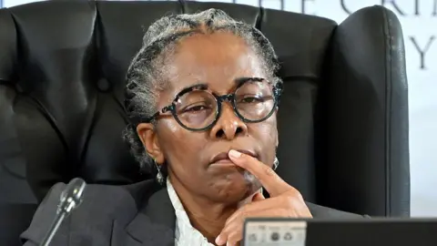 Gallo Images via Getty Images Sesi Baloyi, wearing black framed glasses and a grey suit jacket, holds a finger to her lips