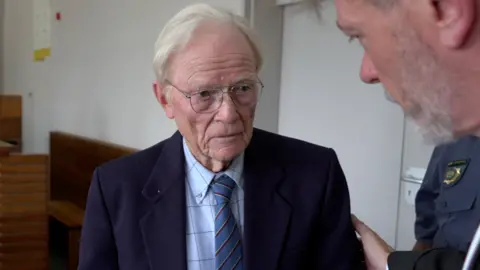 BBC Iain Wares is wearing a navy suit jacket, blue checked shirt and blue tie. He has white hair and glasses. A man is placing a hand on his shoulder as they speak. A court of police officer can be seen in the background.