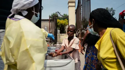 Getty Images A young girl washes her hands at a checkpoint for hand washing and temperature screening for all visitors and patients entering Kyeshero Hospital in Goma, DRC on 18 May 2026. Also pictured is a member of medical staff who wears a gown, gloves and face mask, along with another visitor who also wears a face mask.