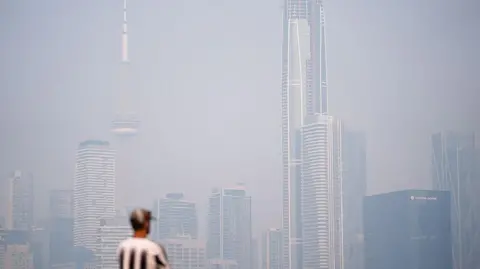 Anadolu via Getty Images A man in a cap and a black and white vertical stripped shirt stands looking at a hazy Toronto skyline
